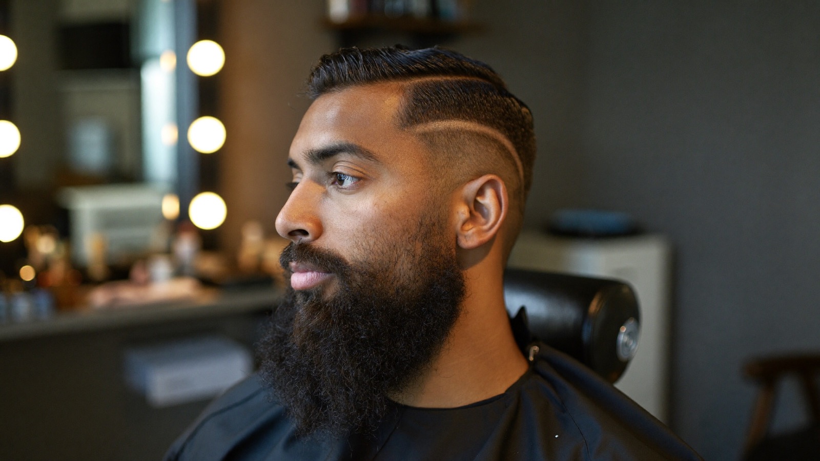 Man in his thirties with a full, groomed dark beard sitting in a barbershop chair under warm light