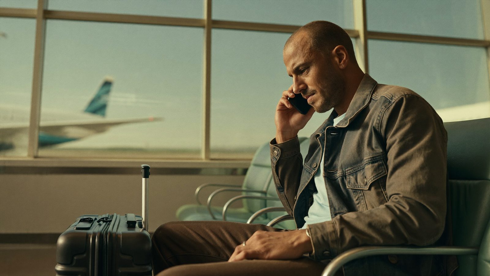 Man with a carry-on suitcase at an airport departure gate looking thoughtfully at his phone