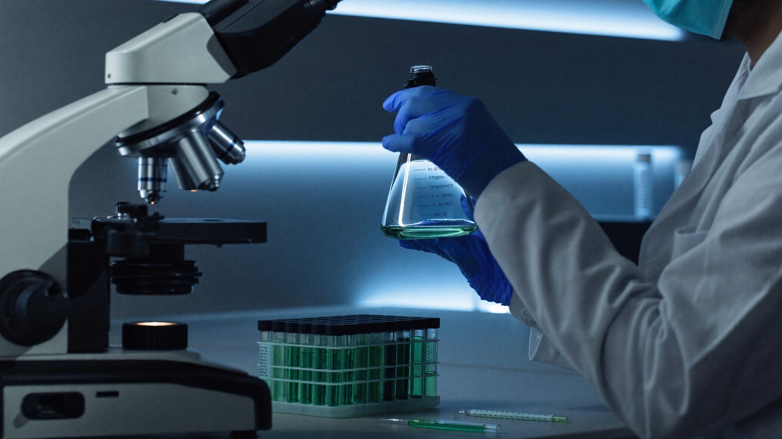 Researcher in a lab coat holding a cell culture flask beside a microscope