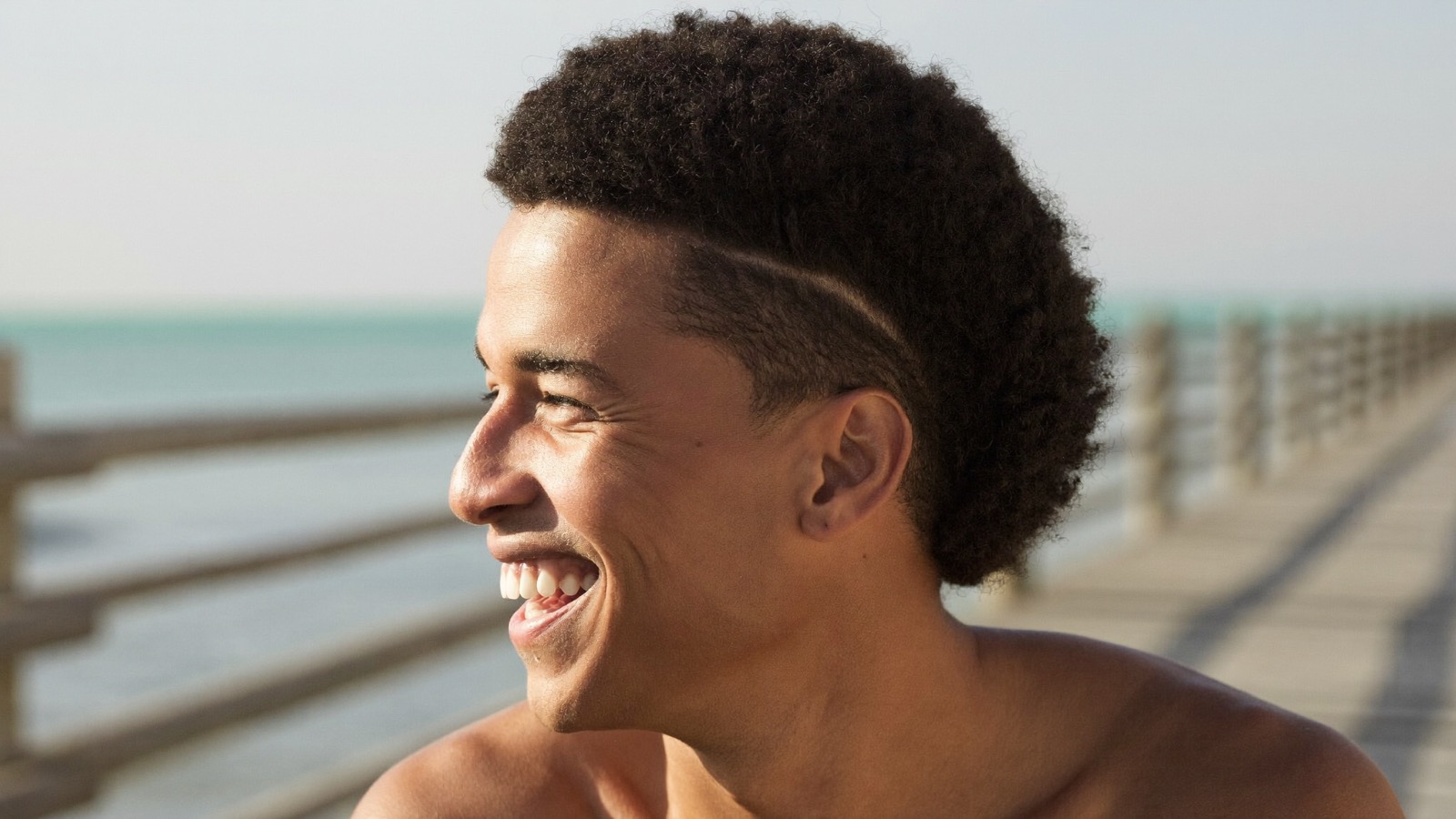Smiling young man with full, curly dark hair on a seaside boardwalk in soft daylight