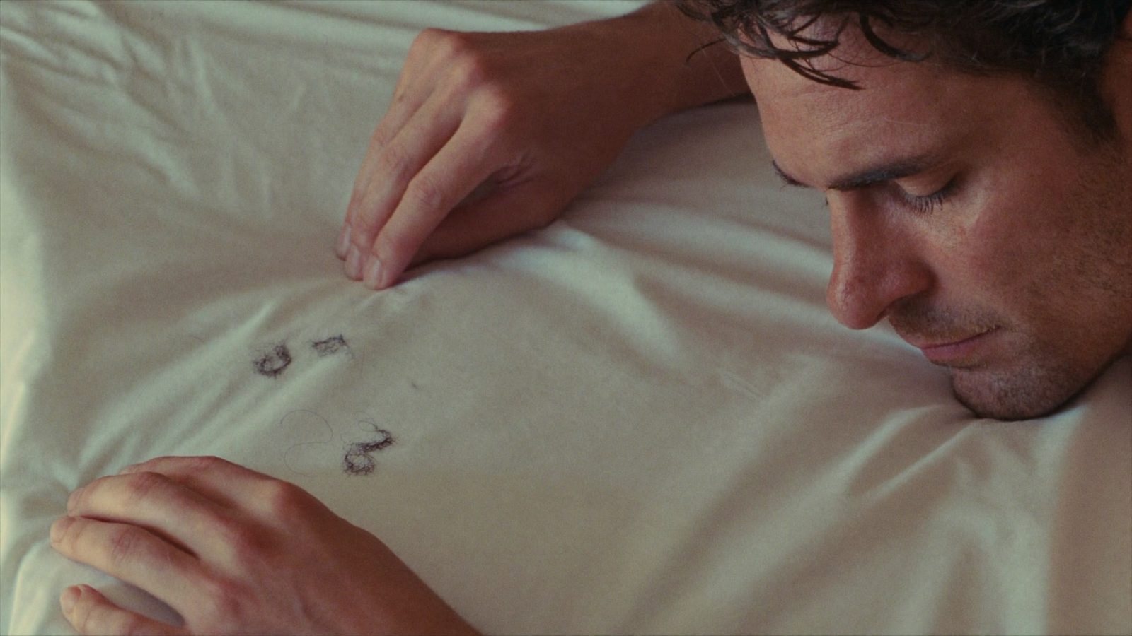 Man in his 30s looking at a few shed hairs on a white pillowcase in soft morning light